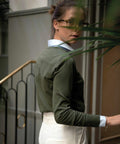 A person wearing glasses, a green sweater over a white collared shirt, and white pants is looking back over their shoulder while standing near a staircase and partially obscured by a Nimbus NB94F.