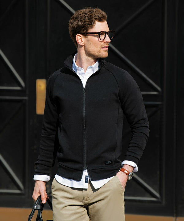 A man wearing glasses and a black zip-up jacket over a white shirt stands in front of a black door, looking to his left. He is holding the Nimbus NB95M in his right hand and wearing khaki pants.