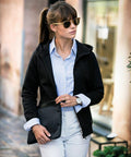 Woman wearing NB96F sunglasses by Nimbus, a black hoodie over a blue button-up shirt, and white pants, holding a black clutch bag while standing outdoors.