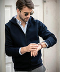 A person wearing sunglasses, a blue hoodie, and a light blue shirt checks their Nimbus NB96M wristwatch while standing against a wall.