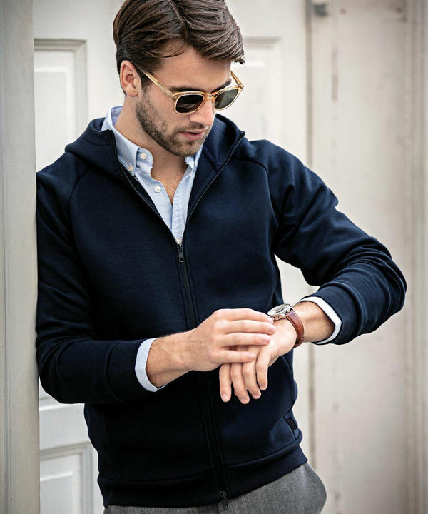 A person wearing sunglasses, a blue hoodie, and a light blue shirt checks their Nimbus NB96M wristwatch while standing against a wall.