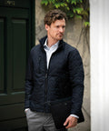 A man with short, wavy hair wears a black NB98M jacket by Nimbus over a white dress shirt and gray pants, holding a black leather folder. He stands in front of a green door with ivy on the wall.