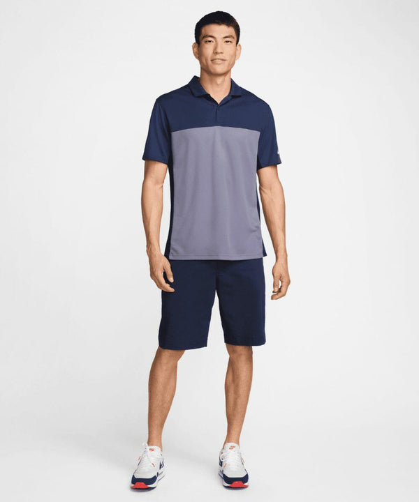 A man is standing against a plain background, wearing a Nike NK170 short-sleeved polo shirt, shorts, and sneakers.