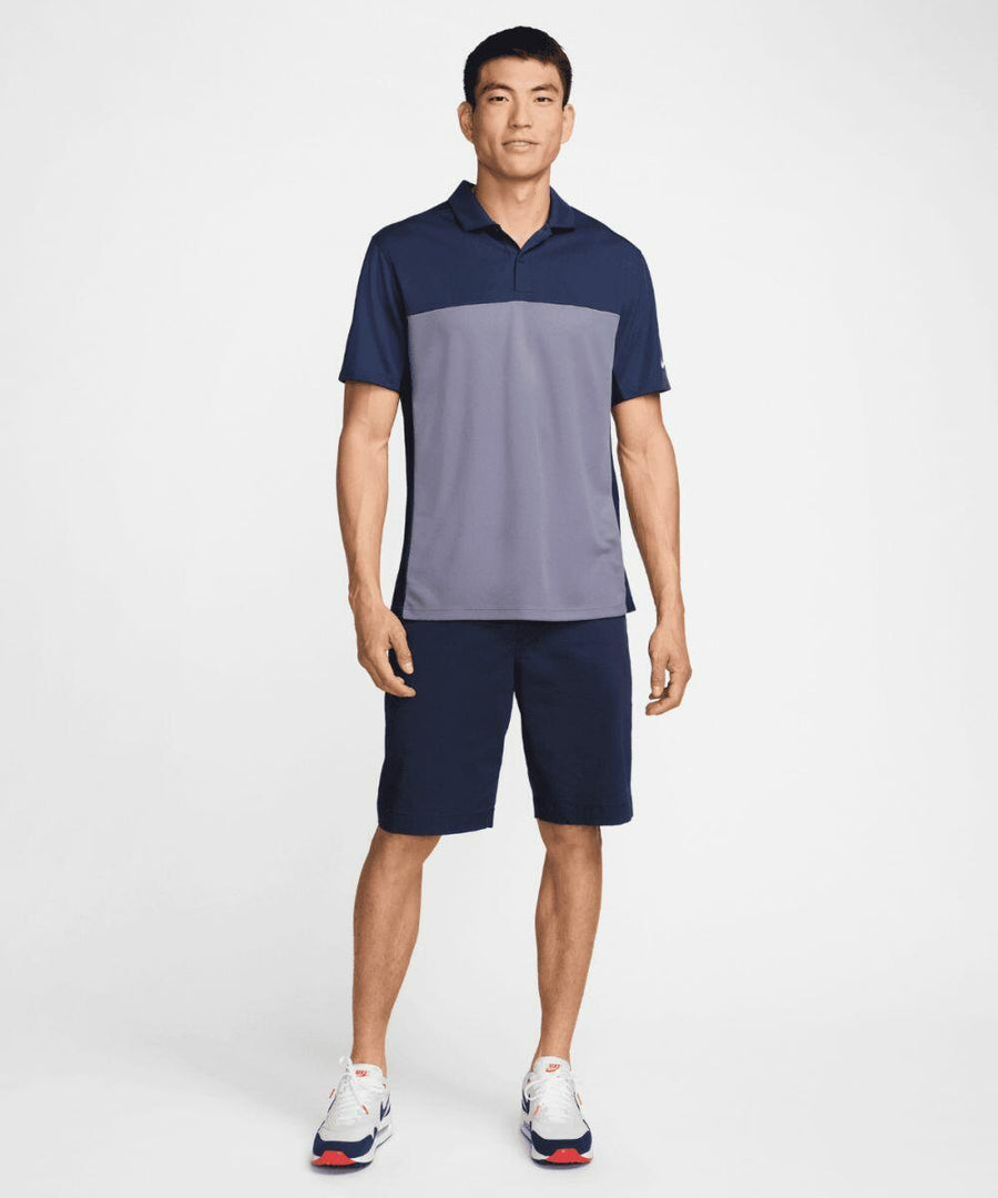 A man is standing against a plain background, wearing a Nike NK170 short-sleeved polo shirt, shorts, and sneakers.