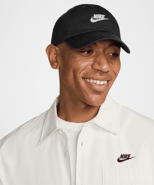 Someone wearing a Nike Club cap in black and a white shirt with the Nike logo smiles whilst looking to the side, against a plain, light-coloured background.