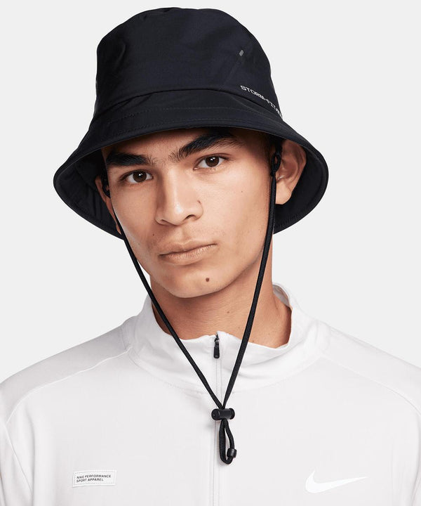 A person wearing a Nike Apex bucket SB hat with chin strap and a white Nike zip-up jacket, posing against a plain light background.