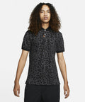Person with curly hair wearing a black, leopard-print Nike NK336 polo shirt and black pants, standing against a plain white background.