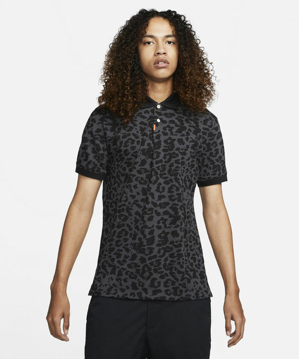 Person with curly hair wearing a black, leopard-print Nike NK336 polo shirt and black pants, standing against a plain white background.