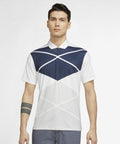 A man stands wearing a Nike NK338 polo shirt with navy and white colors. He has short dark hair and a tattoo on his left forearm. The background is plain white.