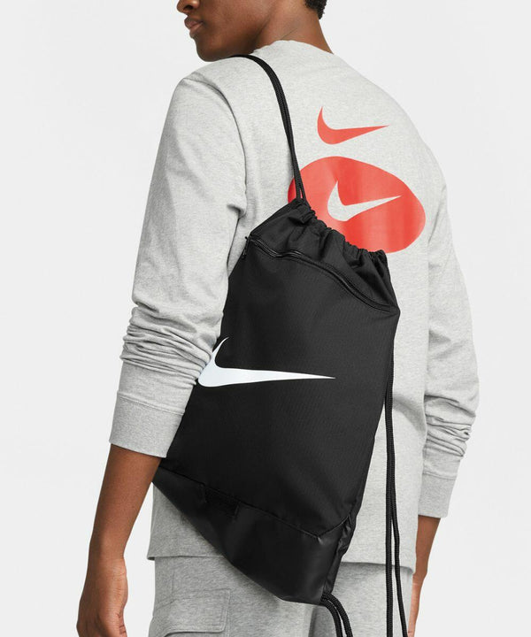 Person wearing a gray Nike sweatshirt with red and white swooshes, carrying a black Nike NK352 drawstring bag with a white swoosh logo.