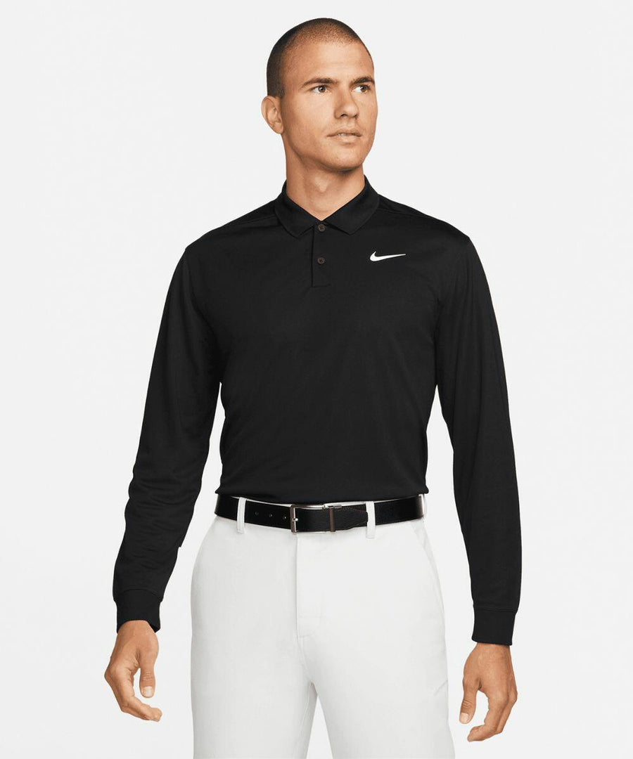 A man is wearing a black Nike NK354 long-sleeve polo shirt and white pants, standing against a plain white background, looking to his left.