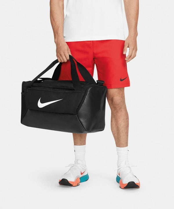 Person in a white shirt and red shorts holding a black Nike NK364 duffel bag; wearing white Nike shoes with teal and orange soles.