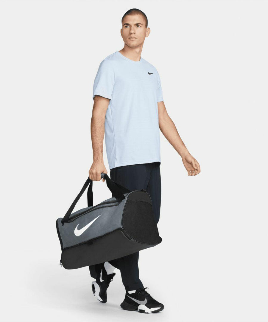 A person wearing a light blue Nike T-shirt, black pants, and white sneakers is carrying a black NK371 Nike duffel bag.