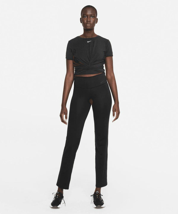 A person stands facing forward wearing a black knotted athletic top, black high-waisted leggings with a front cutout detail, and black Nike NK374 sneakers against a plain white background.
