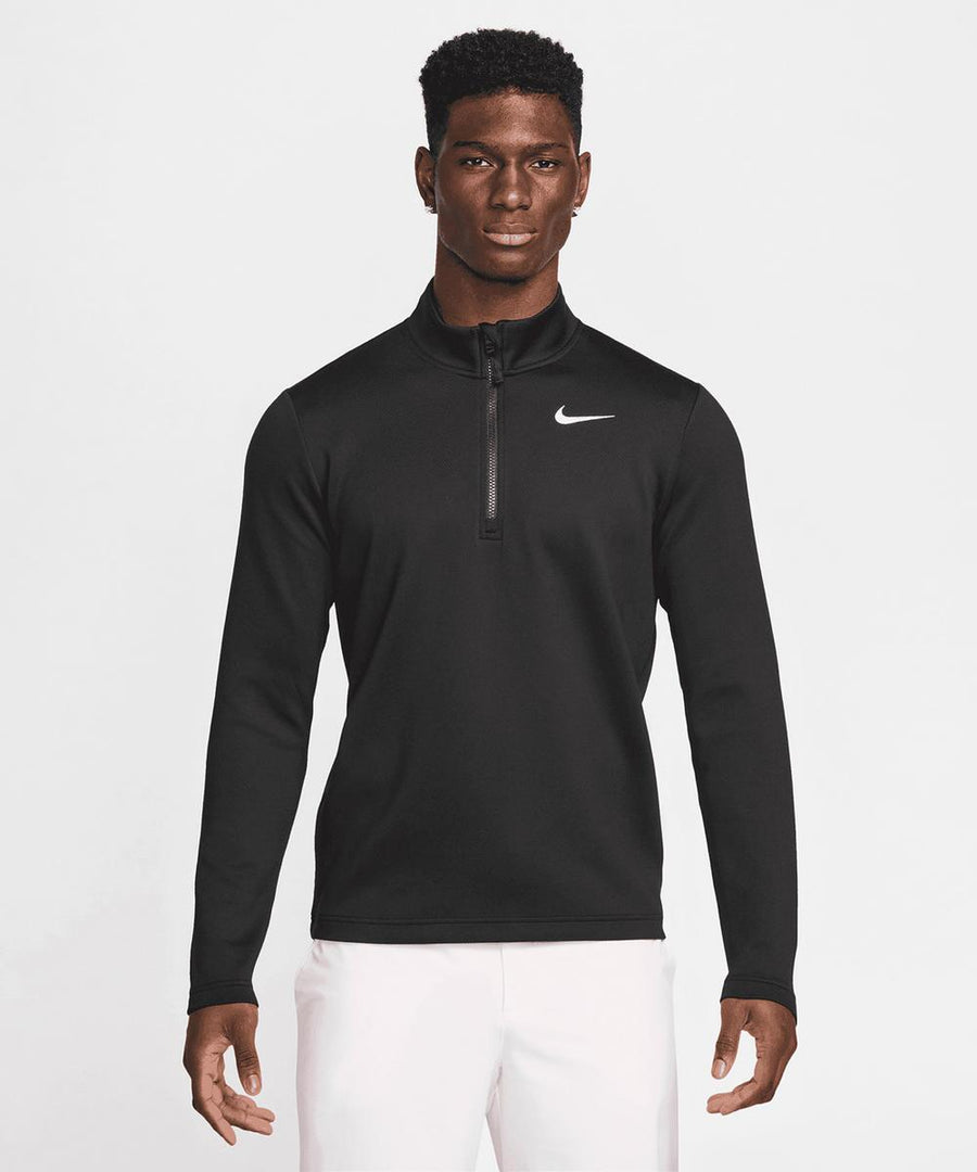 A man faces forward, wearing a black Nike Dri-FIT Victory Therma Flex half-zip top featuring a white Nike logo on the chest, paired with white trousers. The background is plain white.