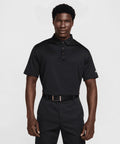 A man stands facing forward, wearing a black Nike Dri-FIT Tour solid polo with a three-button placket and black trousers. His expression is neutral and his arms are relaxed at his sides.