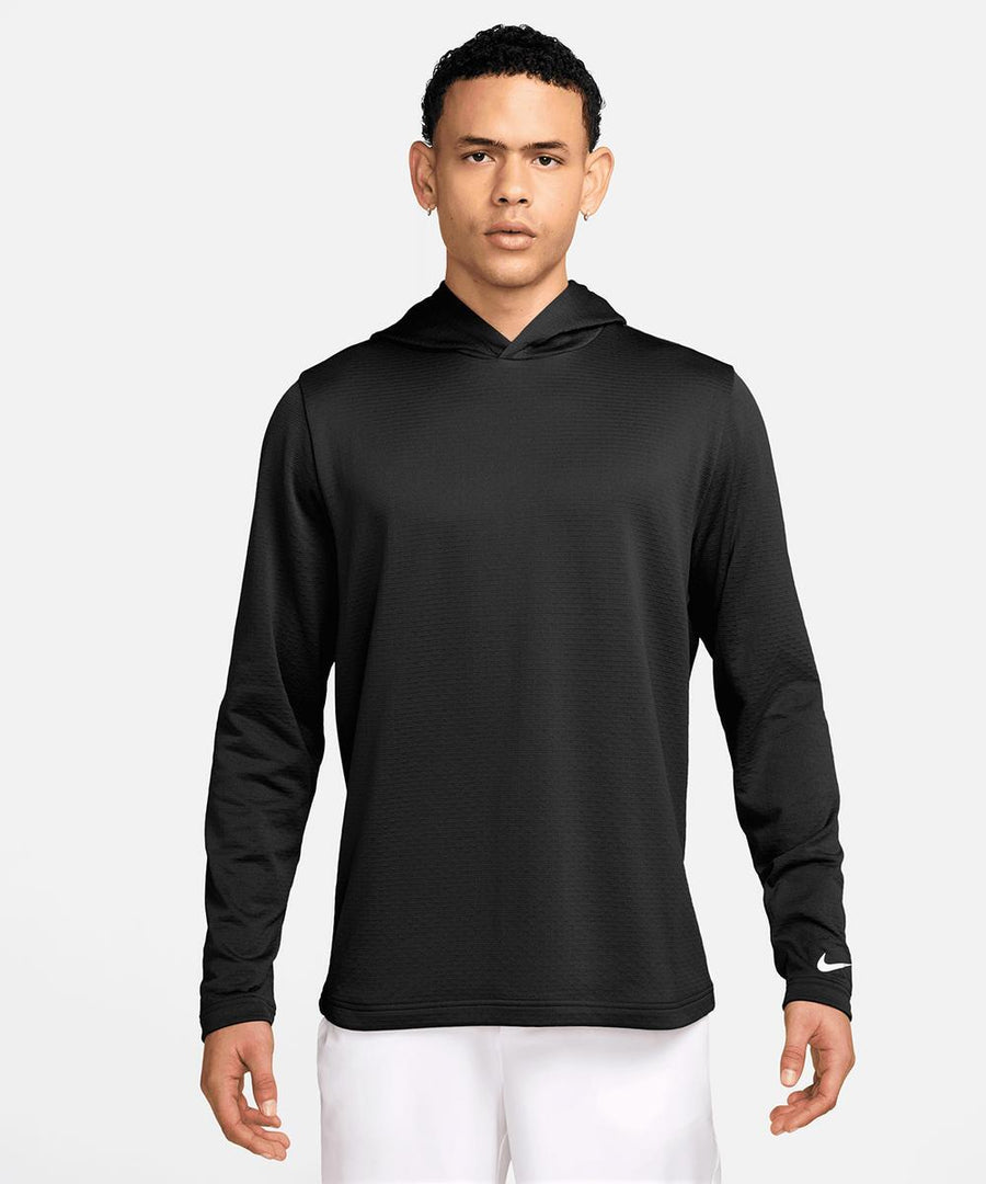 A person faces forwards wearing a Nike Dri-FIT Tour hoodie in black, featuring long sleeves and a small white Nike logo on the left wrist, paired with white trousers against a plain light background.