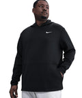 A man stands with one hand in his pocket, looking left, wearing a black Nike Dri-FIT Victory Therma Flex hoodie with a white logo on the left chest and light-coloured trousers. The background is plain white.