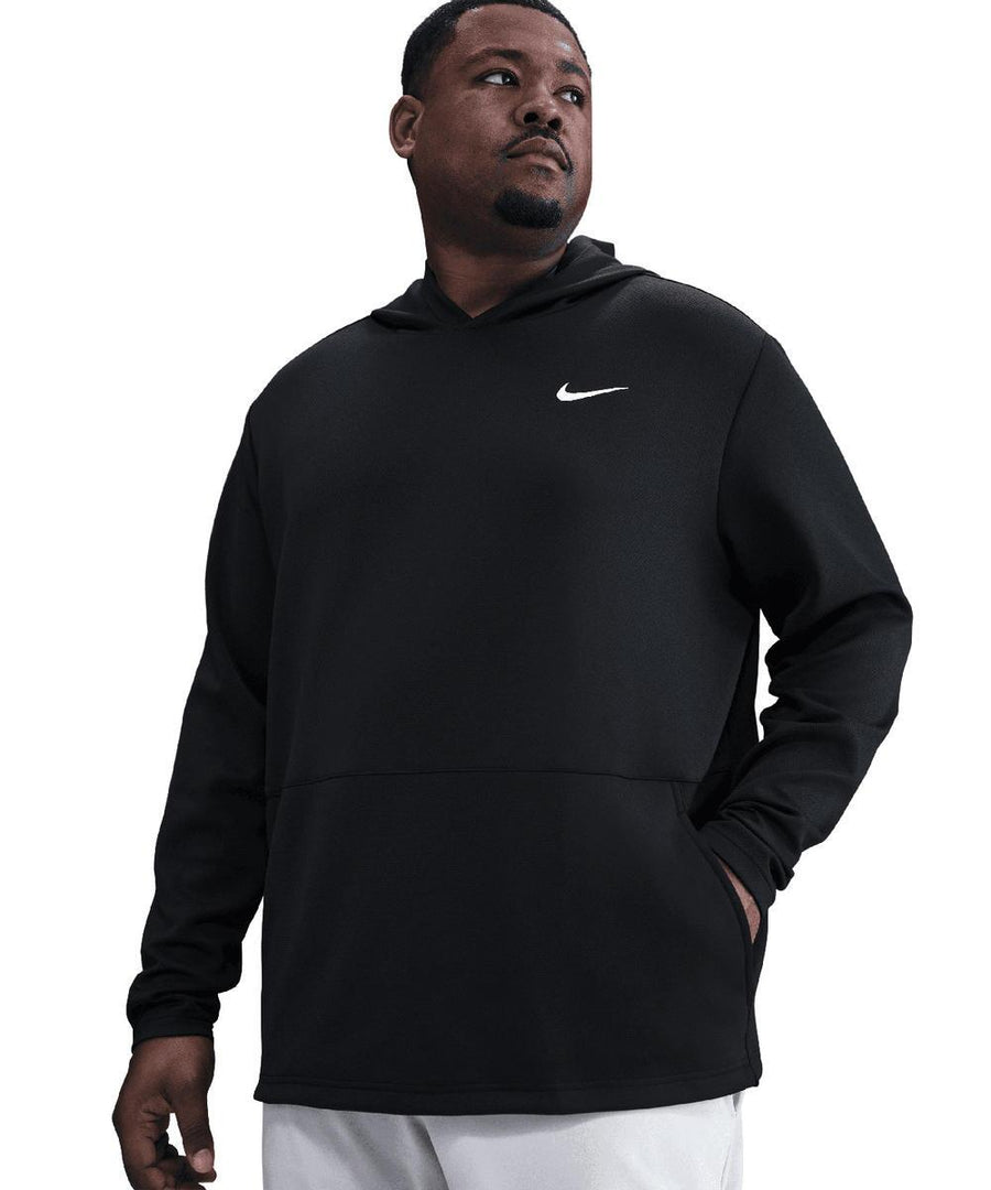 A man stands with one hand in his pocket, looking left, wearing a black Nike Dri-FIT Victory Therma Flex hoodie with a white logo on the left chest and light-coloured trousers. The background is plain white.