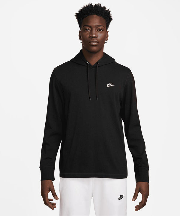 A person stands facing forward in a black Nike Club knitted hoodie, featuring a small white Nike logo on the chest, paired with white trousers with a small black Nike logo. The background is plain white.
