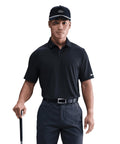 A man in a black Nike Velocity solid polo, black Nike cap, and black trousers stands holding a golf club, facing forwards against a plain white background.