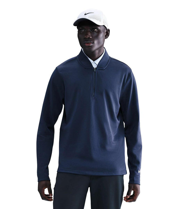A man stands facing forward in a navy Velocity 1/2-zip top by Nike over a white collared shirt, dark trousers, and a white Nike baseball cap. The background is plain white.