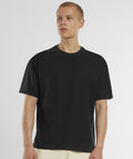 A person with short blonde hair wears the New Morning Studios Oversized Jersey tee in black with light-coloured trousers, standing against a white background and looking slightly to the side.