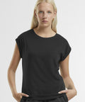 A woman with long blonde hair wears the Women's regular extended shoulder tee by New Morning Studios, standing facing the camera with hands in her pockets against a white background.