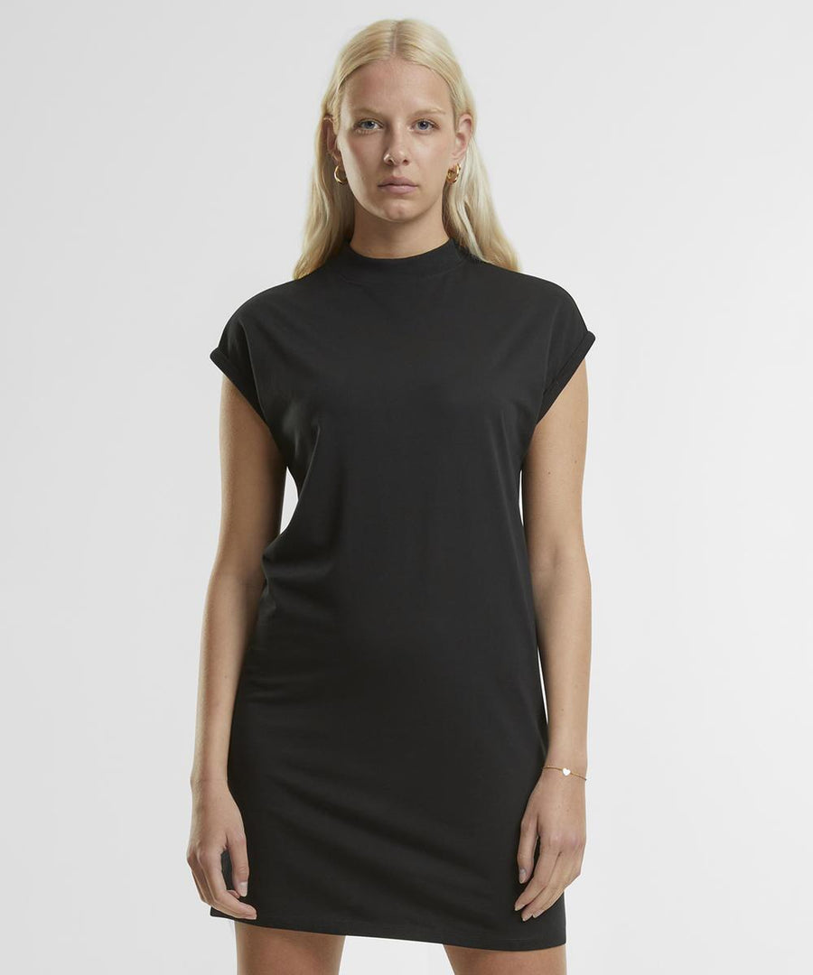 A person with long blonde hair stands against a white background wearing the Women's extended shoulder dress by New Morning Studios, a short-sleeved, black, knee-length outfit. They face the camera with a neutral expression.