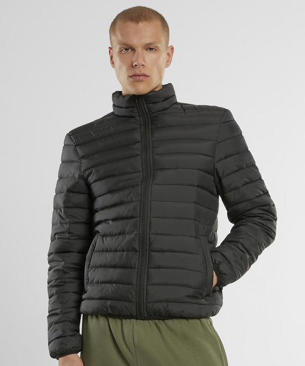 A person wearing the New Morning Studios Light puffer jacket, a black quilted zip-up with horizontal stitching, stands with one hand in their pocket against a plain light background.