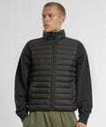 A person models the New Morning Studios Light puffer gilet in black, layered over a black long-sleeve shirt with olive green trousers, standing against a plain white background and gazing slightly to the side.