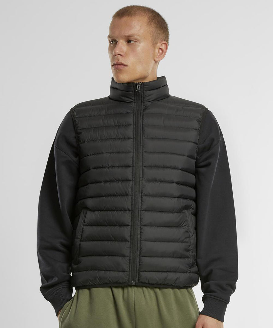 A person models the New Morning Studios Light puffer gilet in black, layered over a black long-sleeve shirt with olive green trousers, standing against a plain white background and gazing slightly to the side.