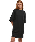 A woman with long, straight, reddish-brown hair wears the New Morning Studios Women's heavy oversized tee dress in black, standing against a plain white background.