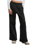 A person models New Morning Studios Women's wide-leg joggers in black with a white long-sleeved top and grey trainers, standing against a plain white background. The photo is cropped from shoulders to feet.