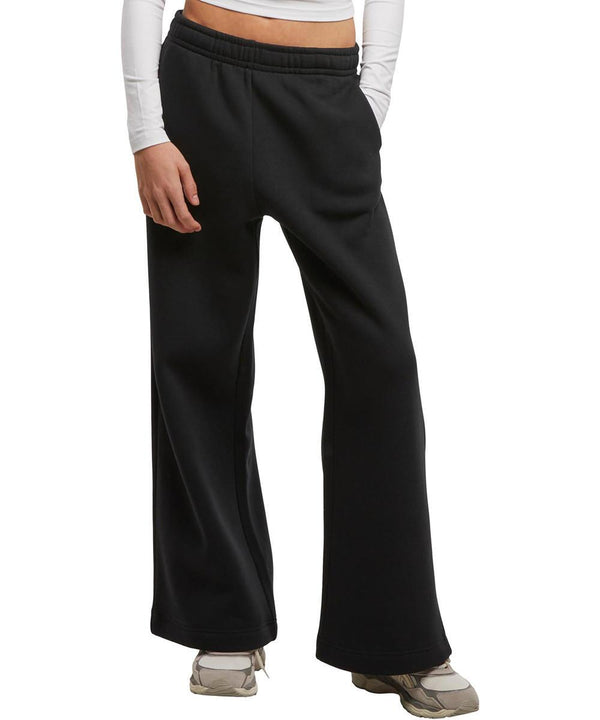 A person models New Morning Studios Women's wide-leg joggers in black with a white long-sleeved top and grey trainers, standing against a plain white background. The photo is cropped from shoulders to feet.