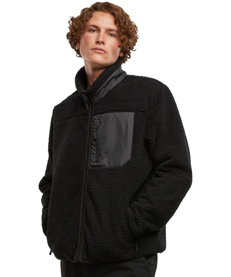 A person with curly hair wears the Bonded Sherpa Jacket by New Morning Studios—a black fleece jacket with a high collar, zip fastening, and black chest pocket. They have their hands in the jacket pockets and gaze slightly upward.