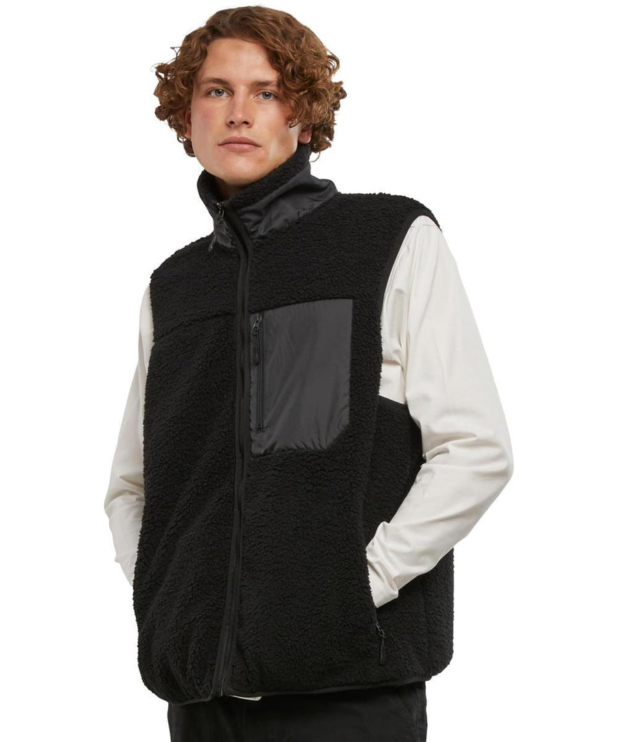 A person stands against a plain background with hands in the pockets of a black Bonded Sherpa Gilet by New Morning Studios, worn over a white long-sleeved shirt, featuring a zip-fastened chest pocket and looking slightly to the side.