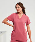 A woman with brown hair wearing an Onna NN310 pink V-neck scrub top and matching pants stands with one hand on her hip against a plain background.