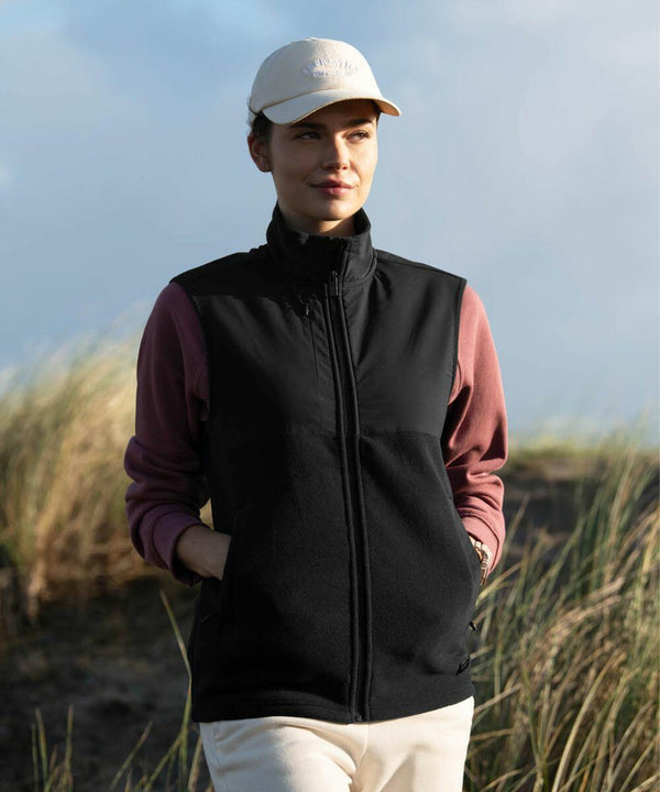 Person outdoors wearing a white cap, black sleeveless Nimbus NP018 jacket, and pink long-sleeve shirt, standing with hands in pockets against a backdrop of grass and cloudy sky.