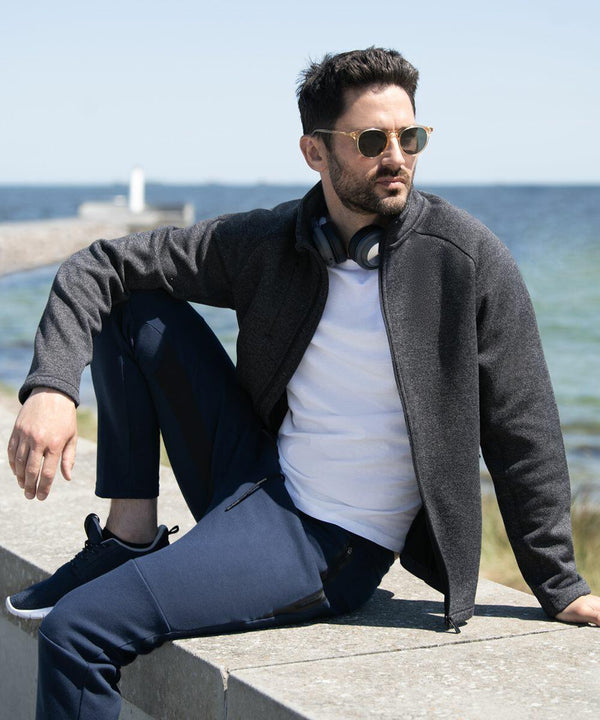 A man with Nimbus NP08M sunglasses and headphones around his neck sits on an outdoor ledge, wearing a gray jacket, white shirt, and blue pants. The background shows a seascape under a clear sky.