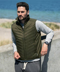 A man wearing a green Nimbus NP10M insulated vest over a gray long-sleeved shirt stands by a concrete wall with a beach and ocean in the background.