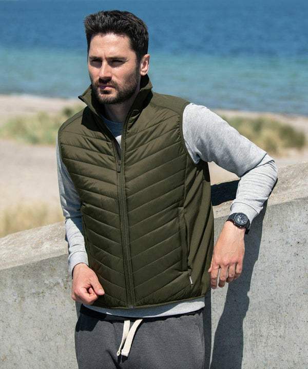A man wearing a green Nimbus NP10M insulated vest over a gray long-sleeved shirt stands by a concrete wall with a beach and ocean in the background.