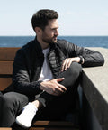 A man in a black jacket and gray pants sits on a wooden bench by the sea, looking to the right, with a Nimbus NP11M beside him.
