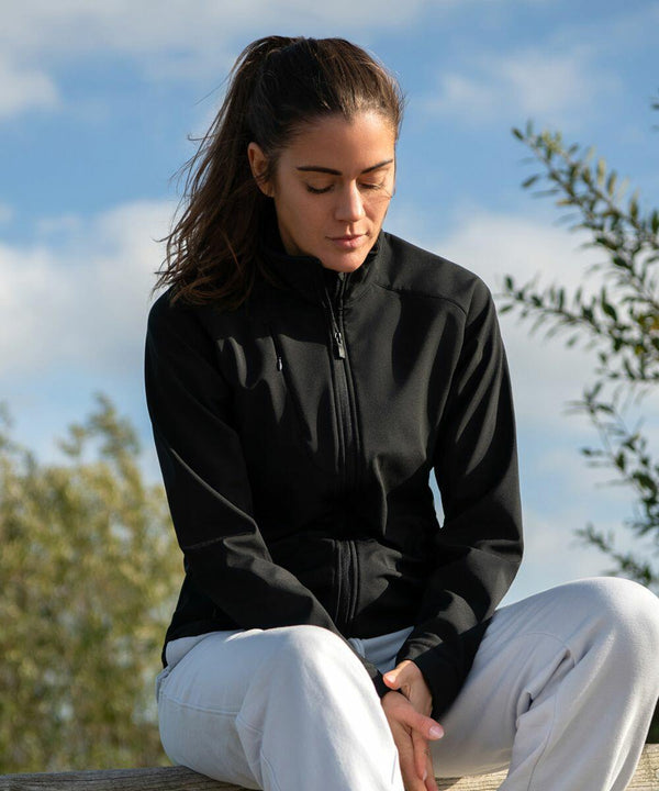 Woman with a ponytail sits outdoors wearing a black Nimbus NP12F zip-up jacket and white pants, looking down with a thoughtful expression. Trees and a cloudy sky are in the background.