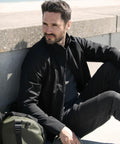A person with short dark hair and a beard, wearing a black jacket and pants, sits on a concrete ledge next to a green Nimbus NP12M backpack, looking into the distance.
