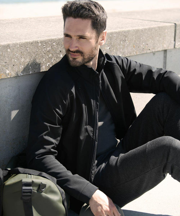 A person with short dark hair and a beard, wearing a black jacket and pants, sits on a concrete ledge next to a green Nimbus NP12M backpack, looking into the distance.