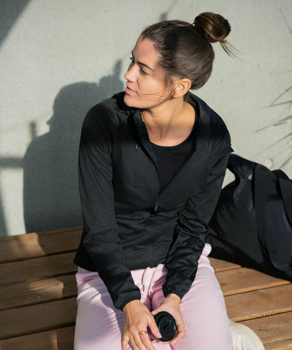 A woman with a bun hairstyle, wearing a black jacket and light pink pants, sits on a wooden bench holding an NP14F by Nimbus. There is a black bag beside her.