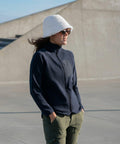 A person wearing a white hat, sunglasses, a Nimbus NP15F navy jacket, and green pants stands outdoors against a concrete backdrop.