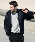 A man in a black jacket and gray shirt is outdoors, holding a Nimbus NP16M bag over his shoulder and looking to the side.