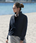 A person wearing Nimbus NP17F sunglasses and a navy jacket stands on a sandy beach with the sea in the background. They have headphones around their neck and hair tied up.